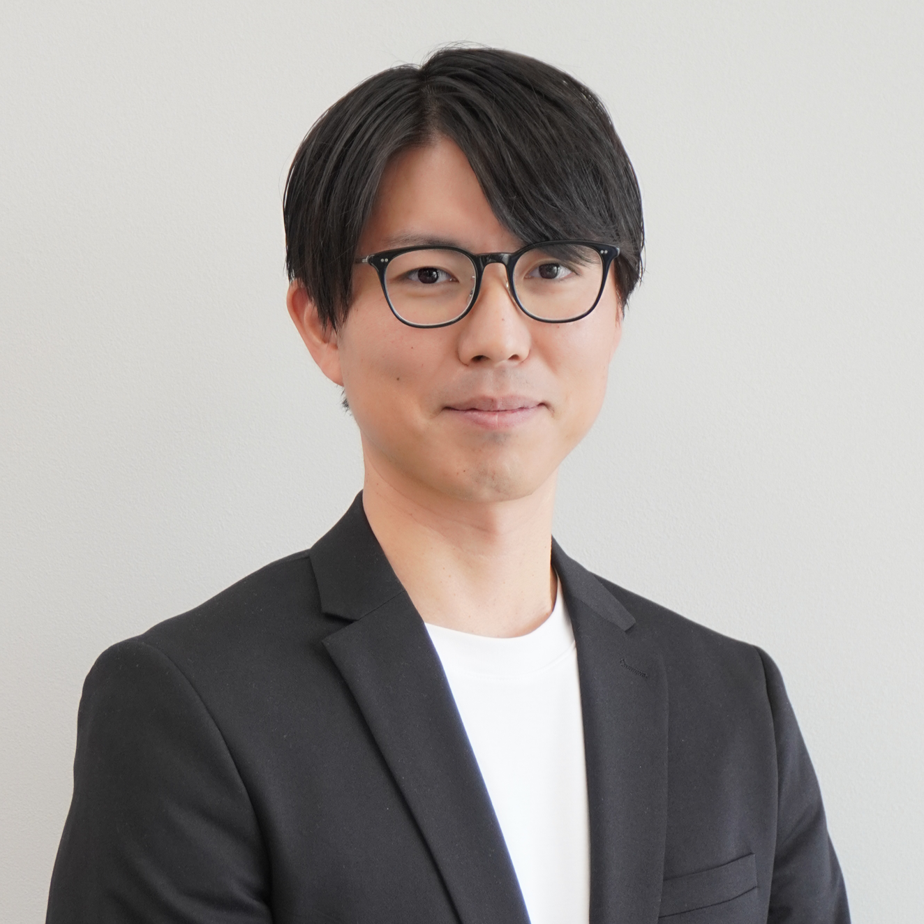 Professional headshot of a man with black hair and glasses, wearing a black blazer and white shirt, neutral background.