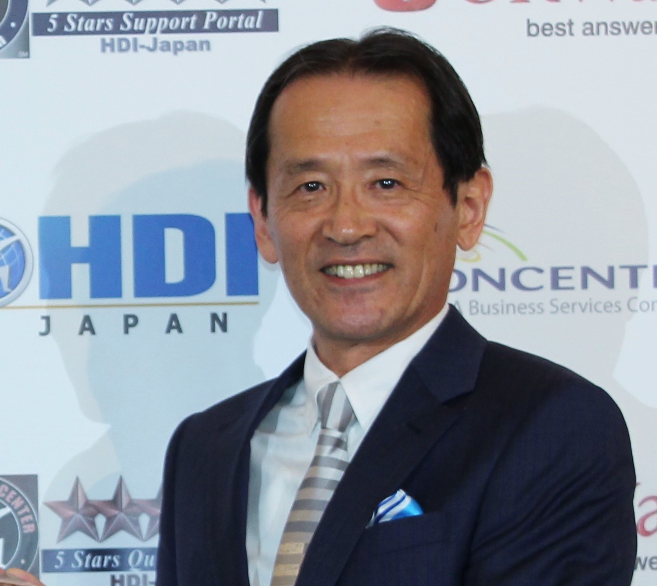 Smiling man in a dark suit with a striped tie posing at a backdrop with logos in the background.