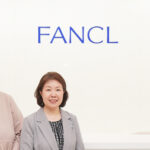 Two women posing in a bright reception area with a FANCL sign on the wall and a MOBILIS logo in the corner behind them.