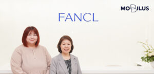 Two women posing in a bright reception area with a FANCL sign on the wall and a MOBILIS logo in the corner behind them.