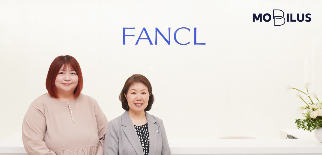 Two women posing in a bright reception area with a FANCL sign on the wall and a MOBILIS logo in the corner behind them.